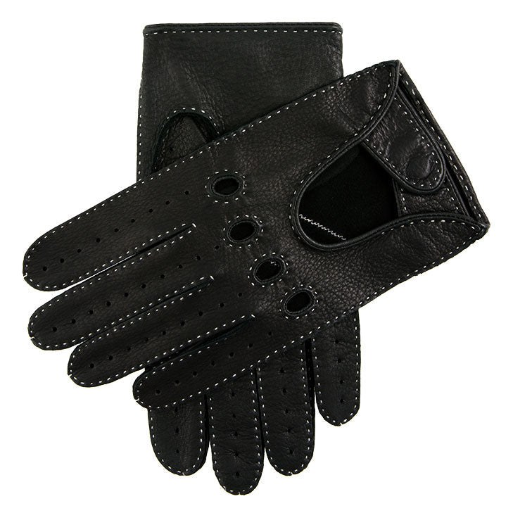 A pair of black handsewn deerskin driving gloves laid flat on a dark surface, cut-out backs with knuckle holes and perforations visible, delicate colour-contrasting stitching catching the light
